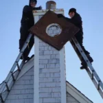 Two people in dark clothing stand on ladders working on a rooftop chimney with a circular metal plate.