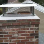 A brick chimney with a metal mesh cap and a rectangular metal cover on top, seen from a rooftop angle.